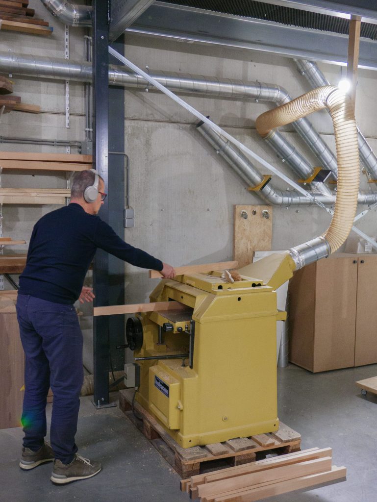 Man werkt aan houtbewerkingsmachine tijdens cursus meubelmaken, met houten planken en moderne apparatuur in een werkplaats gespecialiseerd in houtbewerking.