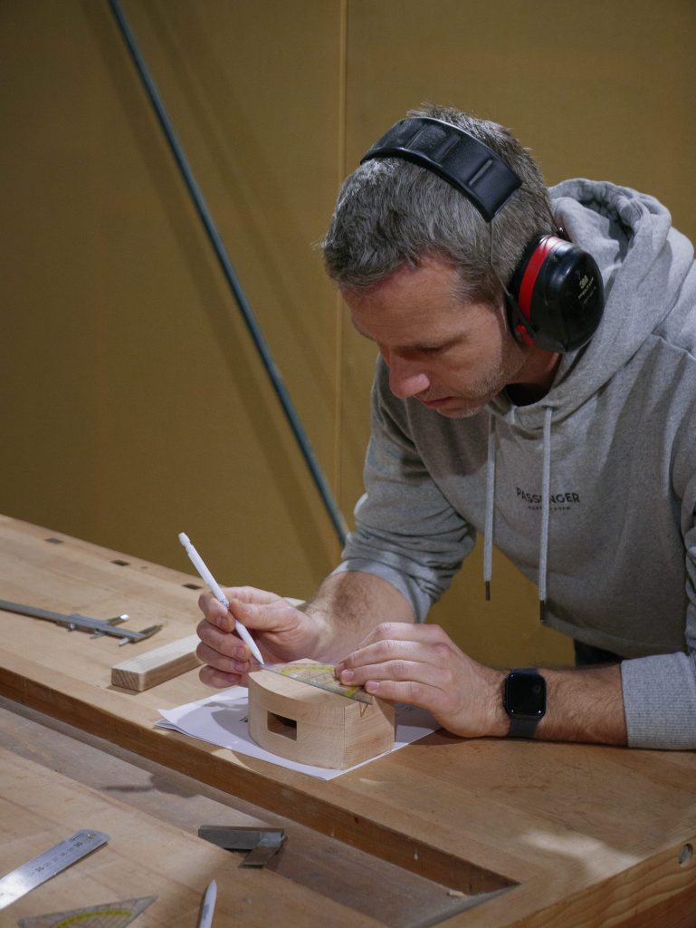 Man volgt cursus meubelmaken en houtbewerking, tekent nauwkeurig op houtblok aan werkbank met gereedschap, draagt gehoorbescherming en hoodie.