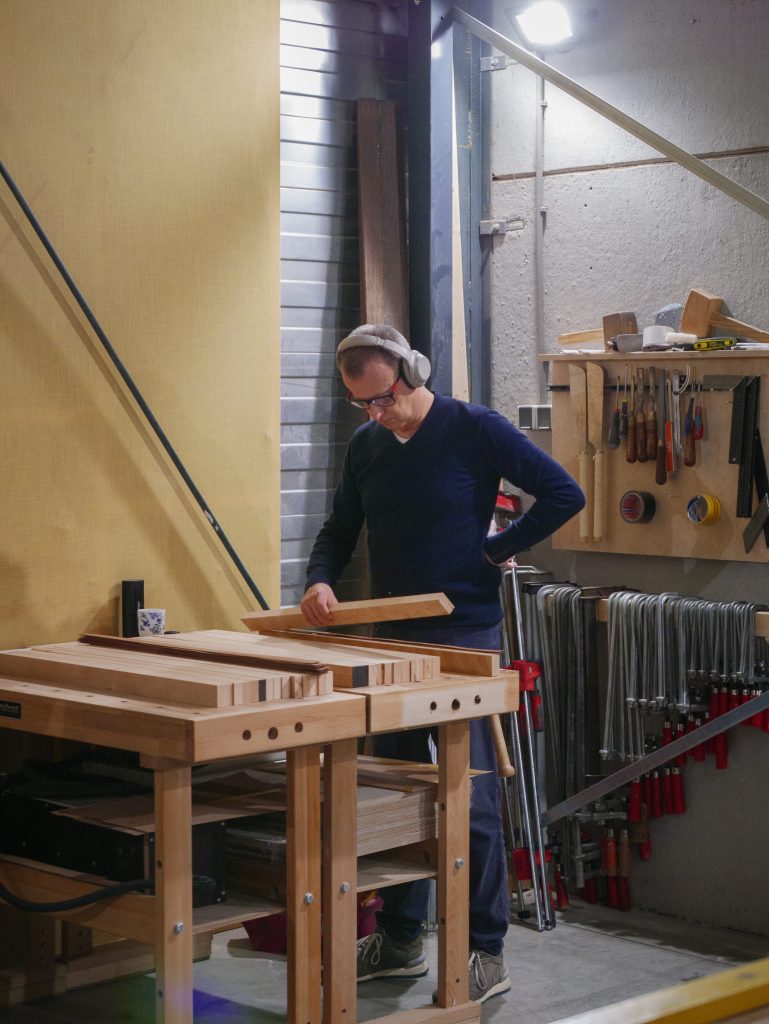 Een man werkt in een houtwerkplaats, draagt gehoorbescherming en onderzoekt houten planken op een werkbank, met gereedschap aan de muur op de achtergrond.