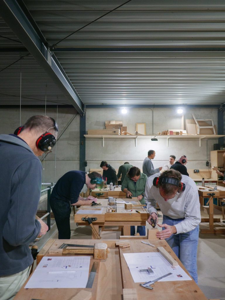 Deelnemers volgen een cursus meubelmaken en houtbewerking in een werkplaats, geconcentreerd bezig met hun projecten en gebruikmakend van verschillende gereedschappen.
