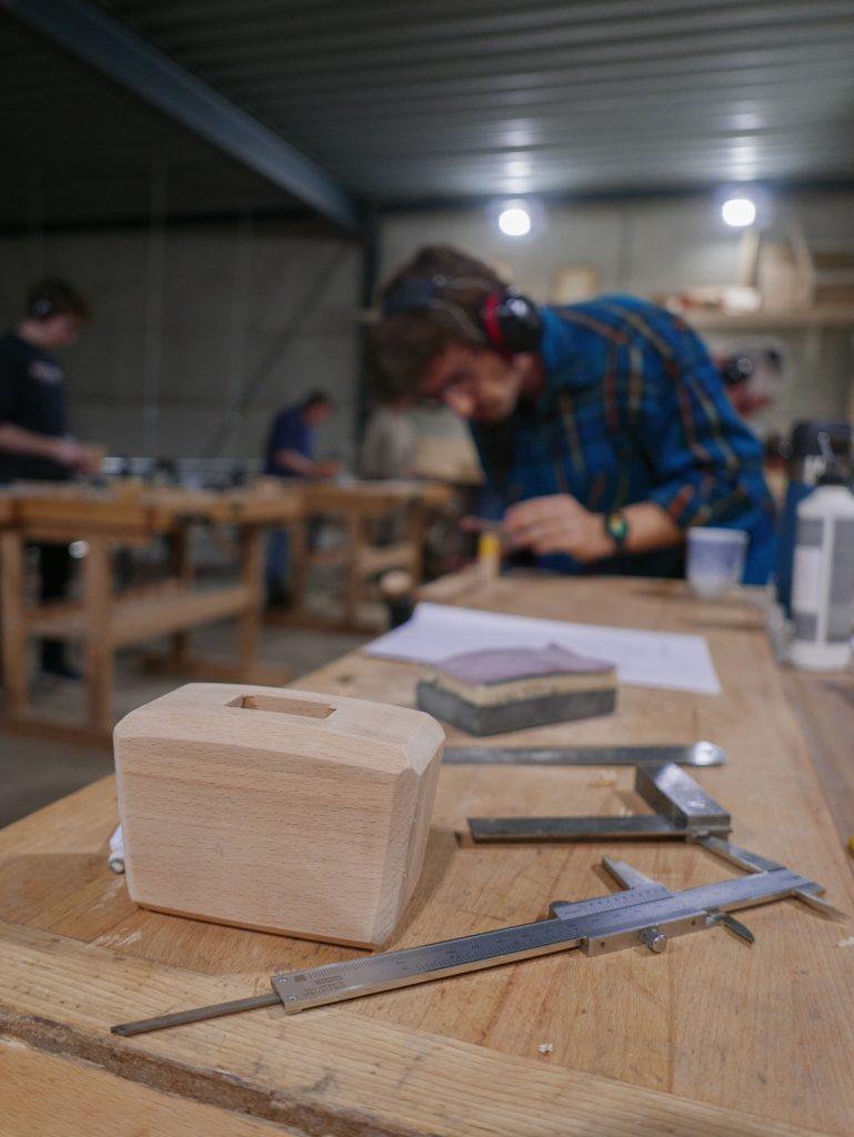 Student volgt een cursus meubelmaken en houtbewerking in een werkplaats, terwijl gereedschappen en houten projecten op de tafel liggen.