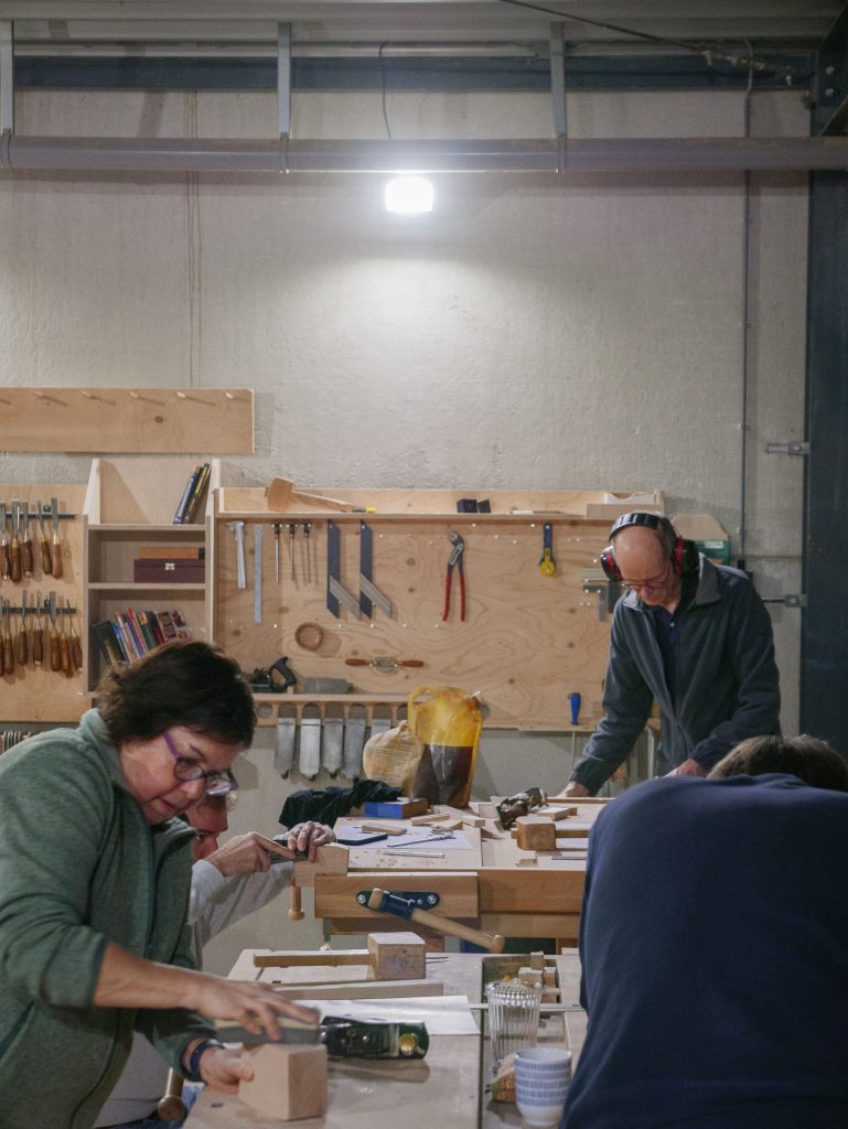Twee mensen werken aan houtbewerking projecten tijdens een cursus meubelmaken in een werkplaats, omringd door gereedschap en houten planken.