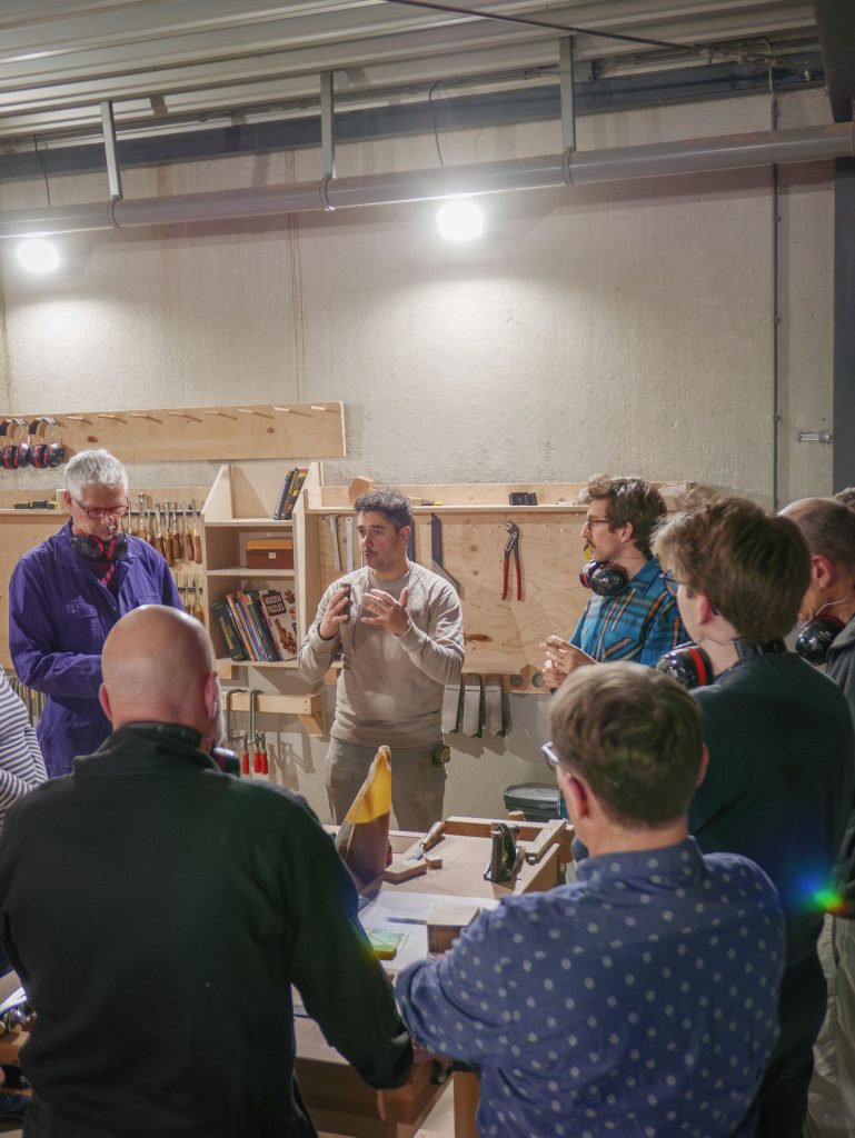 Groep mensen volgt een cursus meubelmaken en houtbewerking in een werkplaats, met aandacht voor ambachtelijke technieken en vakmanschap.