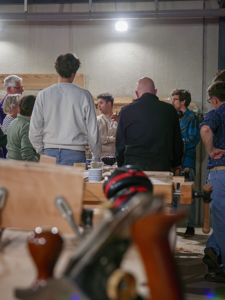 Groep mensen volgt een cursus meubelmaken in een werkplaats, gespecialiseerd in houtbewerking met gereedschappen en houten meubels op de voorgrond.