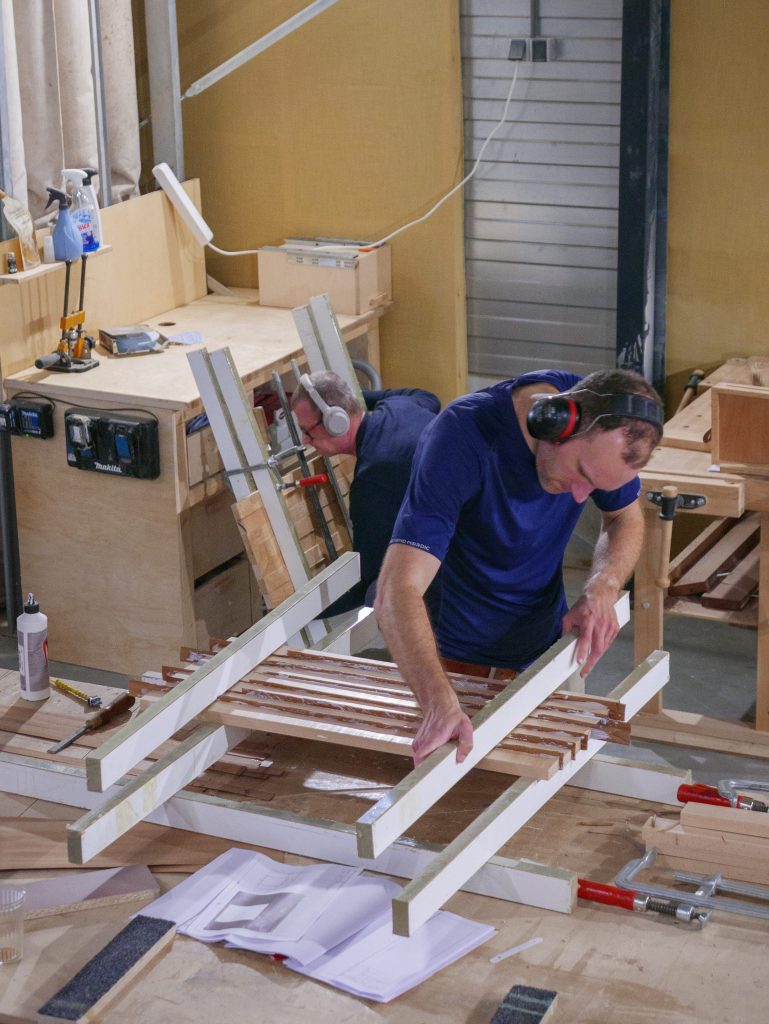 Man bezig met meubelmaken tijdens een cursus houtbewerking, omringd door gereedschap en houten meubels in een werkplaats.