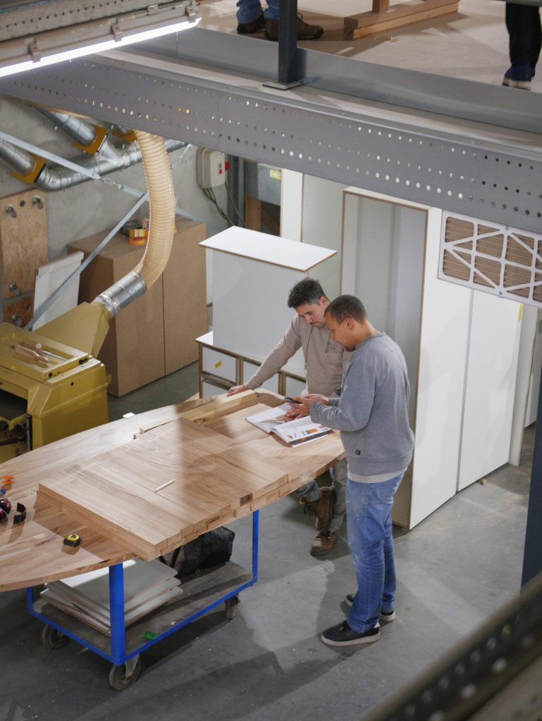 Twee mannen werken samen aan een houten meubel in een werkplaats tijdens een cursus meubelmaken en houtbewerking.