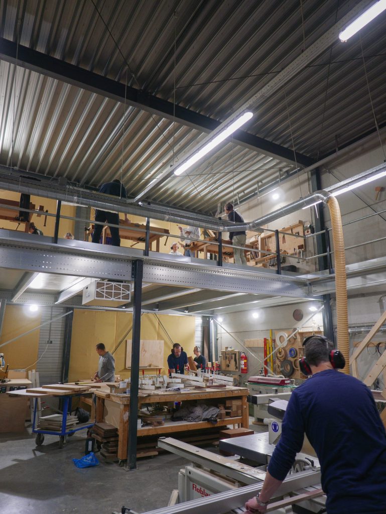 Grote werkplaats waar mensen een cursus meubelmaken en houtbewerking volgen, met meerdere werkbanken en gereedschap in een moderne industriële ruimte.